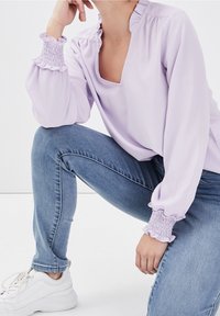Blusa a maniche lunghe color lavanda con collo arricciato e polsini smock. Abbinata a jeans blu e sneakers bianche, posizionata in una posa accovacciata.