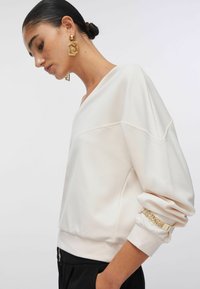 Sweatshirt de couleur crème à coupe décontractée, avec un col large, des poignets côtelés et un détail décoratif en chaîne dorée sur la manche.