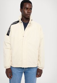 Lys cremet pufferjakke med høj krave, lynlås, elastiske manchetter og sorte brandede detaljer på skulderen. Båret med jeans i blå.