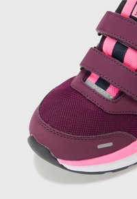 Urheilukenkä, jossa on violetti tekstiilipinta, vaaleanpunaisia yksityiskohtia ja mustia hihnoja. Varustettu teksturoidulla pinnalla ja valkoisella välikohdalla, jossa on harmaa logo.