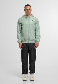 Světle zelená bunda na zip s kapucí, zdobená vyšívaným logem na hrudi, v kombinaci s černými kalhotami a bílými teniskami.