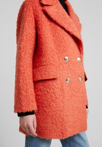 Manteau orange croisé à la finition texturée, boutons en argent, grands revers et poches latérales, associé à un jean bleu.