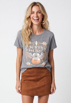 Lachende vrouw met lang blond haar draagt grote hoepels, een grijze "Country Music" t-shirt met gitaarafbeelding en een bruine suède rok.