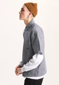 Striped blue and white button-up shirt layered over a gray sweatshirt, paired with a brown knit beanie. Features a chest pocket and logo detail.