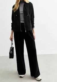 Un hoodie noir zippé, un pull à col roulé gris léger, un pantalon large noir, des baskets blanches et un sac à main matelassé noir, le tout ayant un style décontracté.