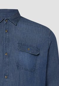 Blaues Jeanshemd mit Knopfleiste, spitzem Kragen und mit Knopf verschlossenem Brusttaschenklappe auf der linken Seite.