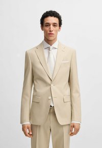Abito beige con un design aderente, giacca a un petto con rever a punta, due bottoni e pantaloni coordinati. Camicia bianca e cravatta inclusi.