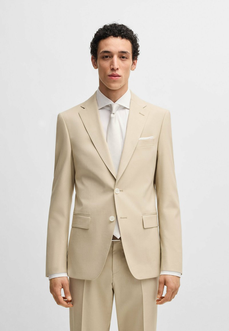 Abito beige con un design aderente, giacca a un petto con rever a punta, due bottoni e pantaloni coordinati. Camicia bianca e cravatta inclusi.