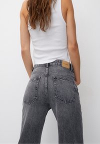 Jean taille haute à jambes larges en denim noir délavé, avec deux poches arrière et un patch logo en cuir tan à la taille.