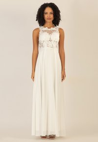 Weißes Kleid mit einem Spitzenoberteil mit floralen Details, transparentem Ausschnitt, fließendem Rock und subtiler Textur, kombiniert mit verzierten Sandalen.