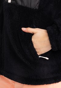 Černá fleecová bunda s jemným, texturovaným povrchem. Obsahuje ruku v přední kapse a bílý zipový detail.