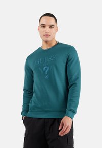 Sudadera teal hecha de una mezcla de algodón, con cuello redondo, mangas largas y un logo bordado con un signo de interrogación en la parte frontal.