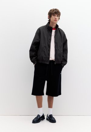 Giovane uomo in piedi indossa una giacca nera con zip, una maglietta a righe, pantaloncini neri fino al ginocchio, calzini bianchi e mocassini in pelle nera su sfondo bianco.