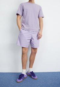 Světlé fialové bavlněné tričko a odpovídající šortky, zkombinované s fialovými semišovými teniskami a bílými ponožkami, na neutrálním pozadí.