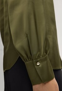 Blusa di raso verde oliva con una texture morbida, caratterizzata da maniche a palloncino e un bottone al polsino per dettagli.