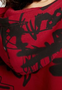 Punainen huppari, jossa on musta abstrakti kuvio selässä. Materiaali näyttää pehmeältä ja siinä on rento istuvuus sekä kiristettävä huppu.