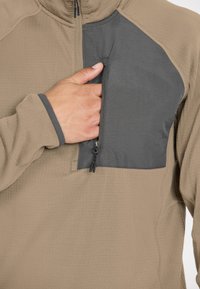 Beiger Fleece-Pullover mit grauem Einsatz und Reißverschlusstasche auf der Brust. Mit geripptem Textil und hohem Kragen. Mit einem Reißverschluss versehen.
