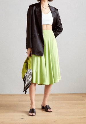 Femme portant un blazer noir, un crop top blanc, une jupe midi vert citron, des chaussures noires à bout ouvert, tenant un sac à main vert avec une écharpe noir et blanc.