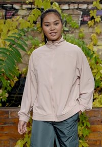 Veste coupe-vent rose clair avec un col haut, fermeture éclair intégrale, poignets élastiques et une coupe décontractée, associée à un pantalon cargo vert.