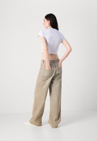 Een vrouw staat met haar rug naar de camera, gekleed in een cropped witte top en wijde beige jeans, met haar handen in haar zakken, tegen een neutrale achtergrond.
