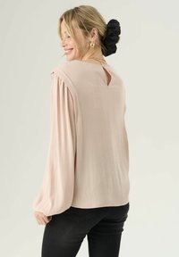 Jasnoróżowa bluzka o długim rękawie z detalem typu keyhole na plecach, z plisowanymi ramionami i teksturowanym materiałem. Połączona z dopasowanymi czarnymi dżinsami.