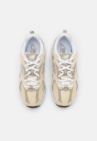 Chaussures de course New Balance beige et blanches avec des lacets blancs, des panneaux en mesh, et le logo "NB" sur les côtés et la languette, vues de dessus.