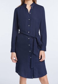 Granatowa sukienka zapinana na guziki, z długimi rękawami, wysokim dekoltem, strukturalnym materiałem oraz związaną w talii paskiem. Posiada pionowe paski i srebrne guziki.
