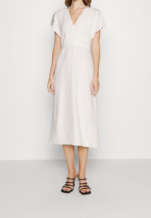 Žena v bílé midi šatech s krátkými rukávy a výstřihem do V, v kombinaci s černými sandály na podpatku s pásky, stojí před jednobarevným pozadím.