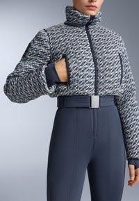 Teksturirana plavo-bijela skraćena jakna s visokim ovratnikom, bočnim džepovima i pritegnutim pojasom. Nosila s tamnoplavim tajicama.