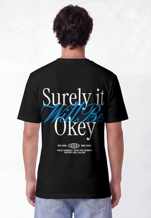 Man draagt een zwart T-shirt met tekst "Surely it will be okay" in wit en blauw, plus datums en een motiverende zin onderaan.