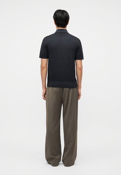 Άνδρας που στέκεται πλάτη, φοράει κοντομάνικη σκοτεινή πλεκτή μπλούζα και φαρδιά καφέ παντελόνια, σε απλό φόντο.