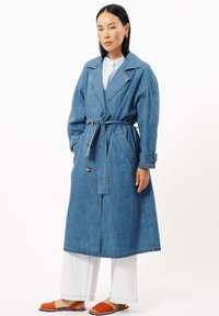 Denim trenchcoat, blauw, met grote kraag, een ceintuur in de taille en zijzakken. Gecombineerd met witte brede broek en oranje sandalen.