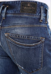 Mörkblå denimjeans som visar bakfickan med brun stickning och en svart rektangulär varumärkeslapp ovanför midjebandet.