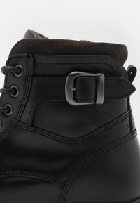Botín de cuero negro con punta redondeada, diseño con cordones, un acento de hebilla en el tobillo y un cuello de ante texturizado.