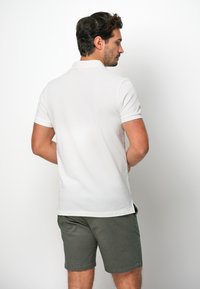 Vit kortärmad polo tröja med platt krage, som har en slim fit och sidofickor, i kombination med olivgröna shorts.