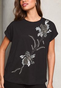 Camiseta negra de manga corta con un diseño intrincado de flores en pedrería plateada y gris en la parte delantera, con un corte holgado y textura suave.