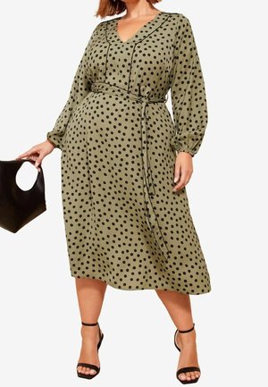 Femme portant une robe midi vert olive à pois noirs, manches longues, col en V et taille nouée, tenant un sac à main noir et portant des talons noirs.