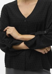 Blusa nera in tessuto texturizzato con scollatura a V, chiusura con bottoni e maniche a volant. Il tessuto presenta piccoli punti in rilievo su tutta la superficie.