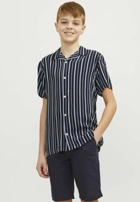 Camisa de manga corta azul marino con rayas verticales blancas, botones al frente y cuello. Combinada con pantalones cortos oscuros, con las manos en los bolsillos.