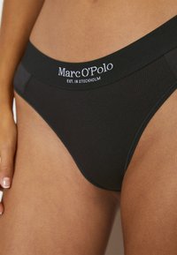 Slip bikini righe nero con un morbido elastico in vita, decorato con il logo bianco "Marc O'Polo" e il dettaglio "EST. IN STOCKHOLM". Texture liscia.