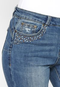 Jeans de denim en un lavado azul medio que cuentan con un diseño decorativo en los bolsillos, con acentos metálicos y un detalle bordado a lo largo de la cintura.