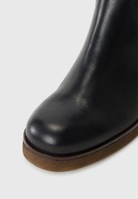 Bottine en cuir noir avec un bout rond, texture lisse et semelle en caoutchouc marron. Elle présente un design élégant pour un enfilage facile.
