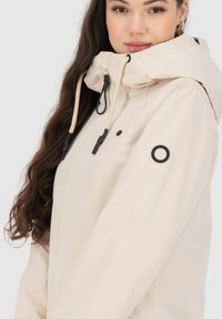 Giacca impermeabile beige con cappuccio, cordoncino e dettagli neri. Presenta una chiusura con zip e un patch con logo sulla manica. Texture liscia.