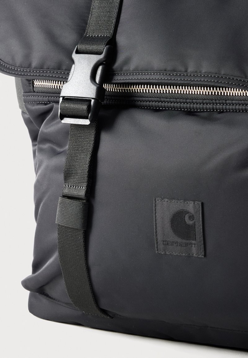 Sac à dos noir Carhartt avec fermeture éclair, sangle réglable et fermeture à boucle en plastique, présentant le logo de la marque sur la poche avant.