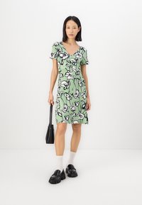 Rochie florală verde cu mâneci scurte, modele din contur negru, tiv până la genunchi, asortată cu mocasini negri și o mică poșetă neagră.