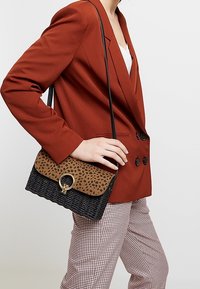 Sac à main tissé noir avec un rabat en imprimé léopard marron, présentant un accent matériel doré circulaire. Le design comprend une longue bandoulière pour le porter à l'épaule.