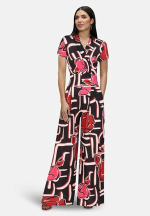 Žena v čiernom overal s širokými nohavicami s ružovými a červenými kvetmi a abstraktnými čiarovými vzormi, stojaca s jednou rukou uvoľnene pozdĺž tela.