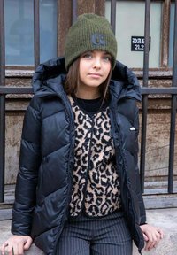 Jeune femme portant un bonnet vert en tricot, une doudoune noire, un pull à imprimé léopard et un pantalon à rayures, appuyée contre une clôture en métal en plein air.