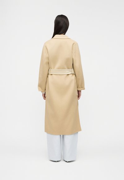 Γυναίκα με μακριά σκούρα μαλλιά, φορώντας μπεζ παλτό με ζώνη και λευκό φαρδύ παντελόνι, στέκεται με την πλάτη στο λευκό, απλό φόντο.