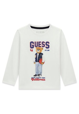 Chemise blanche à manches longues avec un ours en peluche portant une veste universitaire tenant un ballon de basket, texte "GUESS LA EST. 1981 California" sur le devant.